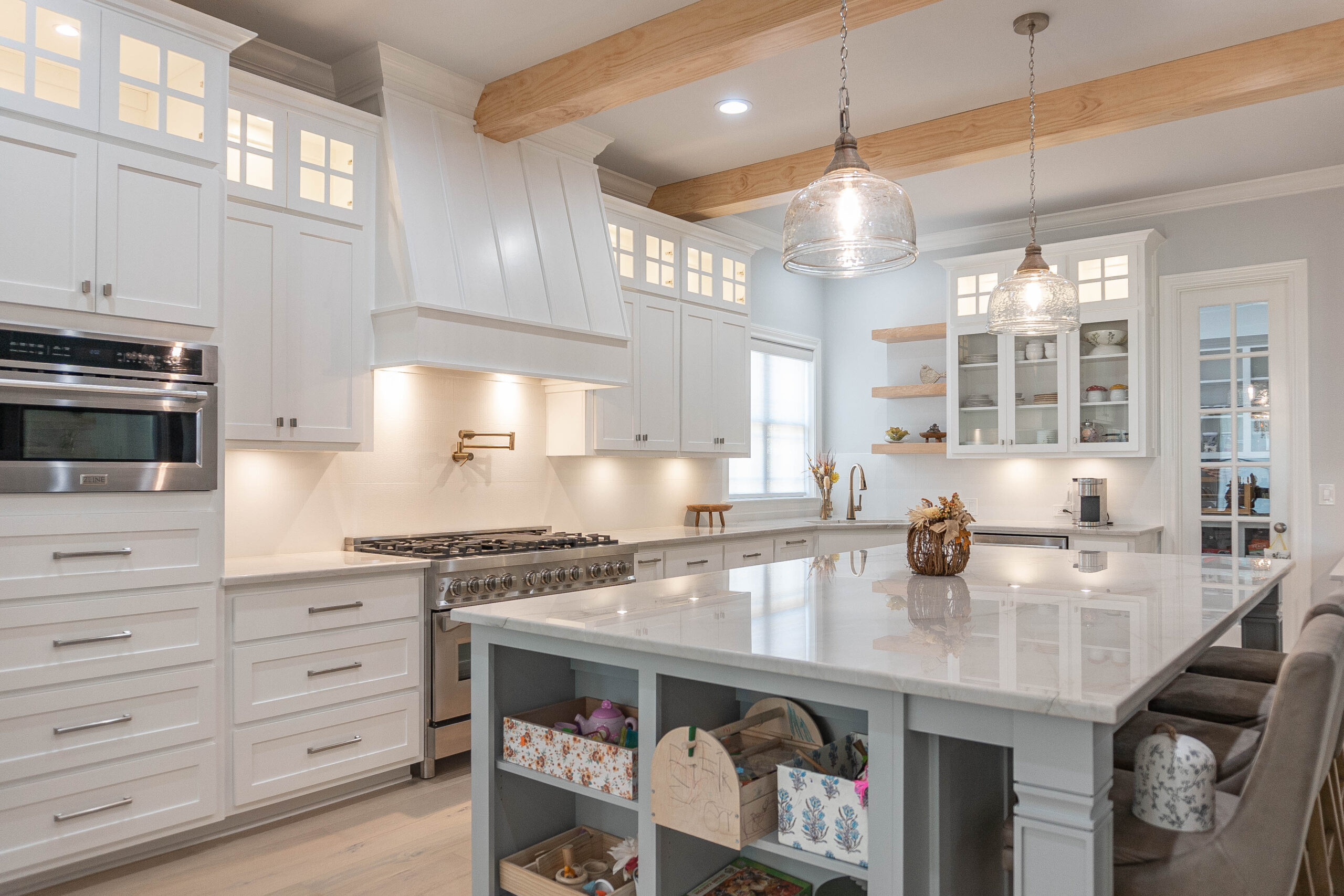 Esplanade Kitchen with natural wood stain ceiling beams and floating shelving with white cabinets glass panel windows tan tile flooring white marble counter top island