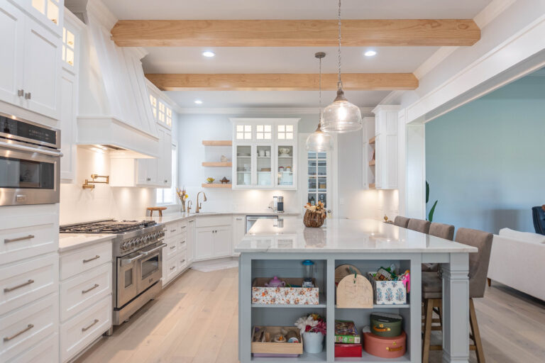 Esplanade Kitchen natural wood finish beam ceiling pendant lights over marble countertop blue island with cut out shelving white tile black splash gold hardware pot filler over gas stove glass cabinet window pantry open to living room Beaumont home builder